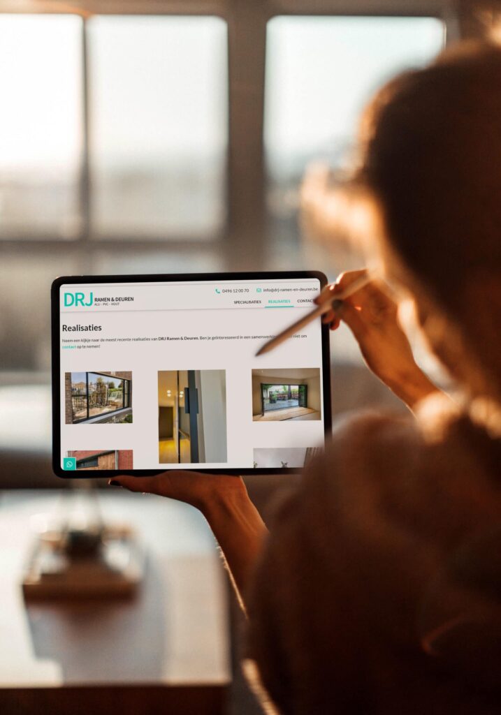 Persoon bekijkt de 'Realisaties' pagina op de tablet met de website van DRJ Ramen & Deuren, met afbeeldingen van hun vakmanschap in raam- en deurinstallaties, tegen een zonovergoten raamachtergrond.