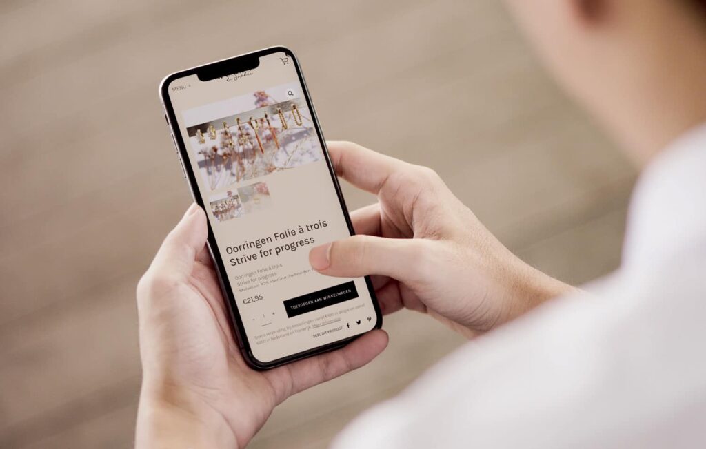 Smartphone in handen van een gebruiker toont de elegante en gebruiksvriendelijke webshop van Tresors de Sophie, met een productpagina van 'Oorringen Folie à trois' weergegeven tegen een neutrale achtergrond.
